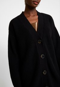 Black knitted cardigan with a textured rib pattern, featuring a v-neckline and three large black buttons on the front.
