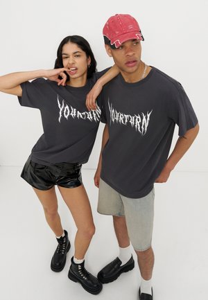 Mlada žena i muškarac nose identične tamne majice s grafičkim uzorkom, žena u crnim kratkim hlačama i čizmama, muškarac u svijetlim kratkim hlačama i crvenoj kapici, poziraju zajedno.