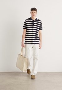 Marineblå og hvite stripete polo-skjorte, hvite bukser, brune slip-on sko, holder en beige tote bag, minimal bakgrunn.