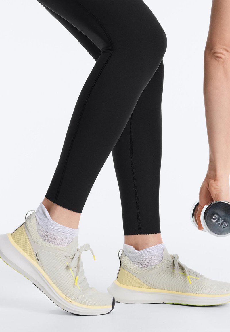 Personne portant des leggings noirs et des baskets jaune pâle soulève un haltère de 4 kilogrammes d'une main sur fond blanc.