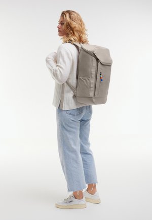 Vrouw draagt een lichtgrijze trui, lichtblauwe cropped spijkerbroek, witte sneakers, met een grote beige rugzak met voorvak met rits.