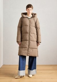 Manteau matelassé marron avec capuche, fermeture éclair sur le devant et manches longues. Porté sur un jean bleu à jambes larges avec un ourlet effiloché. Texture lisse, design chaud.