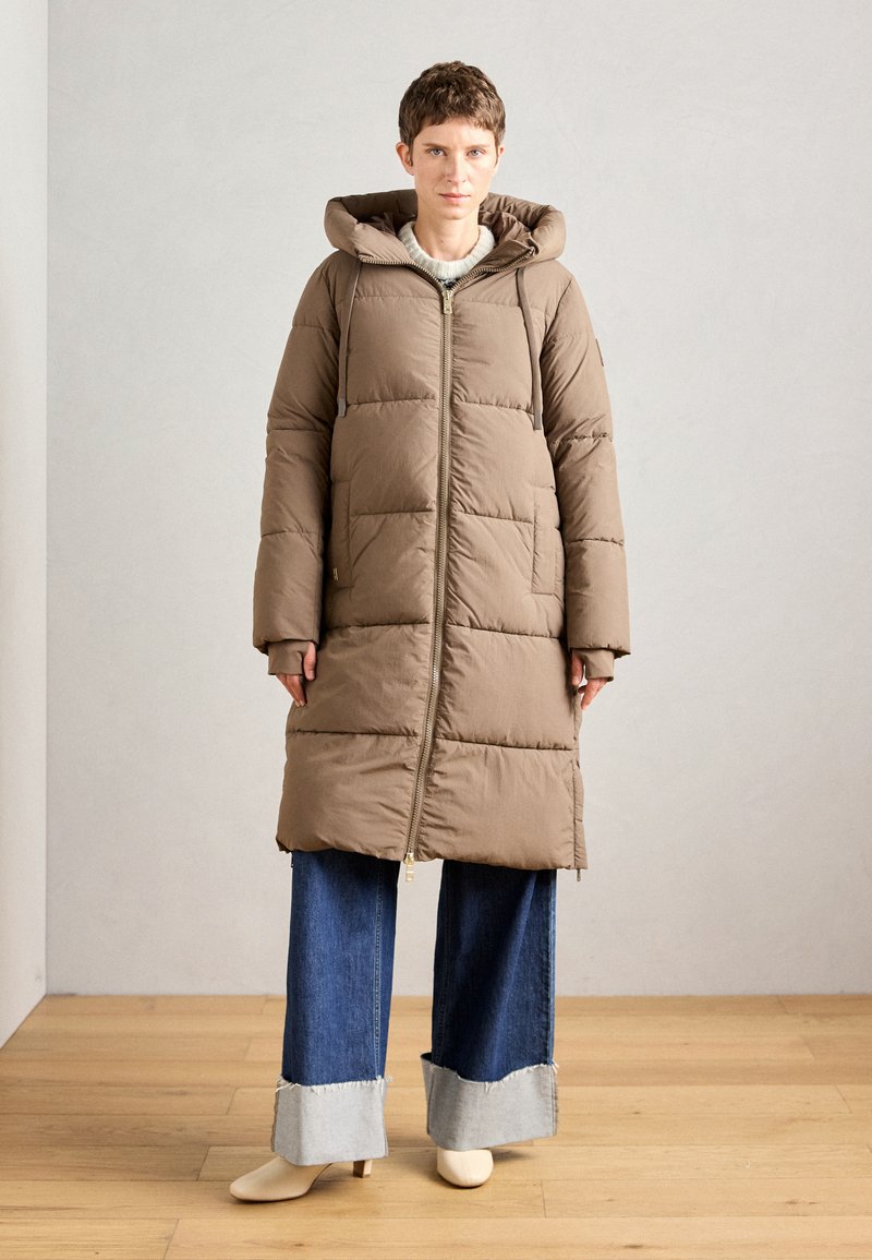 Manteau matelassé marron avec capuche, fermeture éclair sur le devant et manches longues. Porté sur un jean bleu à jambes larges avec un ourlet effiloché. Texture lisse, design chaud.