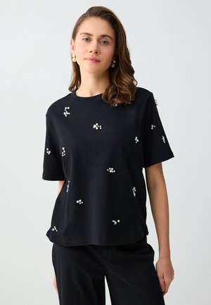 Jonge vrouw met schouderlang bruin haar die een zwart topje met korte mouwen draagt, versierd met kleine parelclusters en bijpassende pareloorbellen.