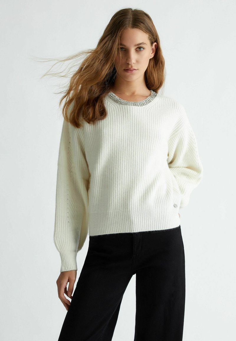 Maglione bianco a coste con scollatura rotonda, impreziosito da perline argento. Vestibilità ampia con maniche lunghe e texture a maglia. Abbinato a pantaloni neri.