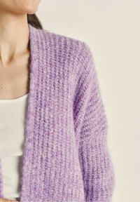 Fialový pletený cardigan s texturovaným vzorem, s otevřeným předním designem a dlouhými rukávy, vrstvený přes světlou tílko.