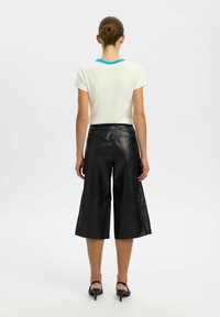 Chemise blanche à manches courtes avec bordure bleue au niveau de l'encolure, associée à un pantalon large en cuir noir taille haute, avec poches apparentes.