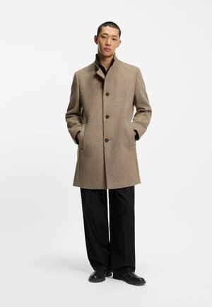 Mand iført beige uldjakke med tre sorte knapper, sorte bukser og sorte sko, stående med hænderne i jakkelommerne mod hvid baggrund.