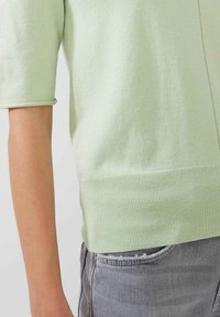 Pull en maille vert clair à manches courtes, tissu texturé, ourlet côtelé, porté sur un denim gris présentant des détails distressed sur la poche.