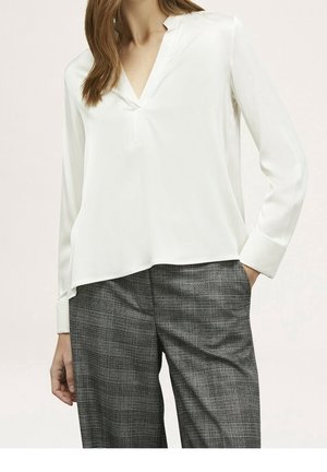 Frau in einer weißen Langarm-V-Ausschnitt-Bluse und grauen karierten Hosen mit einer Hand in der Tasche vor einem hellen Hintergrund.
