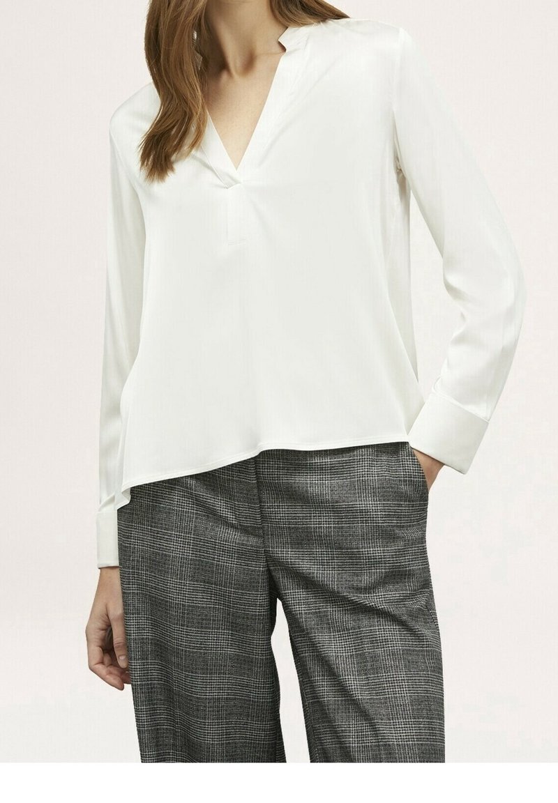 Kobieta ubrana w białą bluzkę z długim rękawem i dekoltem w serek oraz szare spodnie w kratę, trzymając rękę w kieszeni na jasnym tle.
