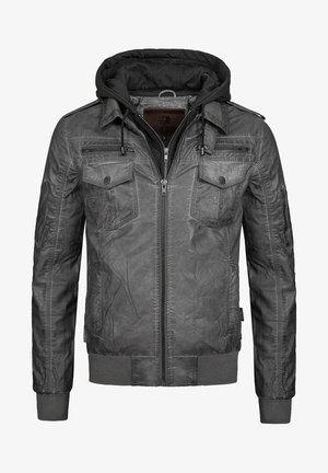 Mann trägt eine graue Lederjacke über einem dunkelgrauen Hoodie, weißem T-Shirt, schwarzen Jogginghosen und weißen Turnschuhen, steht mit einer Hand in der Tasche.