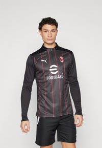 Maglietta da football nera a maniche lunghe con strisce rosse, logo Puma sul petto, colletto con cerniera e la scritta "Football" stampata sul davanti. Indossata con pantaloni corti neri.