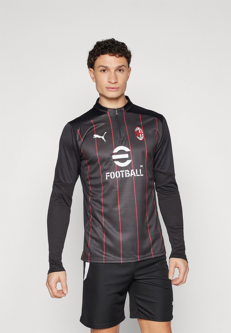Maglietta da football nera a maniche lunghe con strisce rosse, logo Puma sul petto, colletto con cerniera e la scritta "Football" stampata sul davanti. Indossata con pantaloni corti neri.