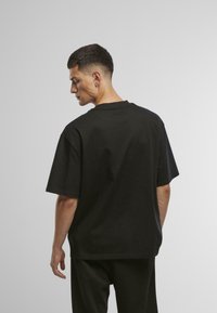 Juodas medvilninis marškinėlis trumpomis rankovėmis ir apvaliu kaklu, su laisvu siluetu ir vientisa dizainu, matomas iš nugaros.