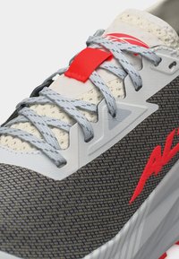 Κοντινή λήψη γκρι αθλητικού παπουτσιού με ανοιχτό γκρι κορδόνια, ύφασμα από δίχτυ, λευκή σόλα και κόκκινο λογότυπο και λεπτομέρειες στην ετικέτα των κορδονιών.