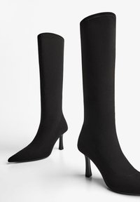 Para czarnych butów do kolan z szpiczastymi czubkami i smukłymi, średniej wysokości obcasami na białym tle.