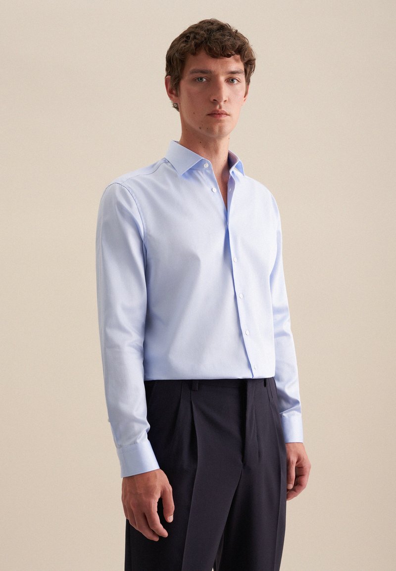 Chemise bleu clair à manches longues avec un col structuré, devant à boutons et texture en coton lisse. Assortie à un pantalon sombre taillé.