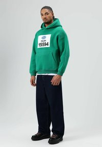 Groen sweatshirt met capuchon met "POLIZEI 15594" gedrukt op een witte patch, gecombineerd met donkere spijkerbroek en zwarte schoenen met roze veters.