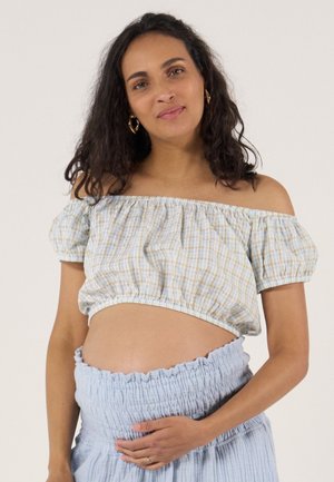 Femme enceinte aux cheveux bouclés, portant un haut épaules dénudées et une jupe, tenant doucement son ventre avec une main.