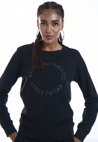 Felpa nera con un design grafico rotondo che riporta la scritta "ESSERE AUTENTICI STREETWEAR" in un testo verde scuro a contrasto.