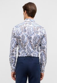 Witte lange mouwen blouse met een blauw en bruin bloemen- en paisleypatroon, voorzien van een klassieke kraag en knoopmanchetten. Donkerblauwe broek zichtbaar.