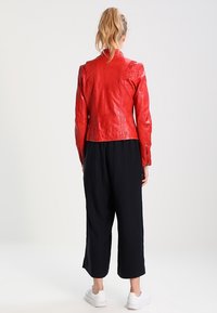 Czerwona skórzana kurtka o kroju crop z dopasowanym fasonem, z wysokim kołnierzem i teksturowanymi ramionami, zestawiona z czarnymi szerokimi spodniami i białymi trampkami.
