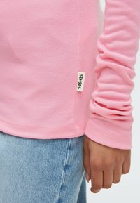 Long-sleeve pink shirt with ribbed texture and a side tag labeled "SENSES." Paired with light blue denim, showing a relaxed fit.