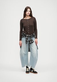 Haut à manches longues marron avec détail de boutons, jeans larges bleu clair avec ourlets effilochés, et chemise à carreaux noir et blanc nouée à la taille.