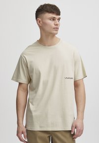 Beige bomulds t-shirt med korte ærmer, rund hals og en lille sort logoaccent nær kanten. Blød tekstur, afslappet pasform.