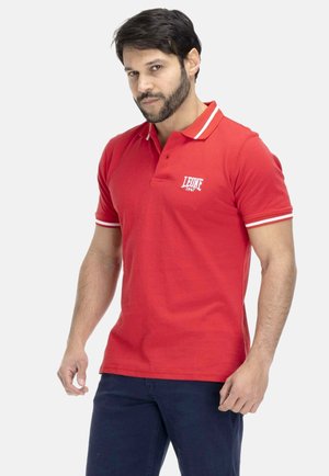 Homme portant un polo rouge à manches courtes avec une bordure blanche et le logo « LEONE 1947 », associé à un pantalon bleu foncé, debout sur un fond blanc.
