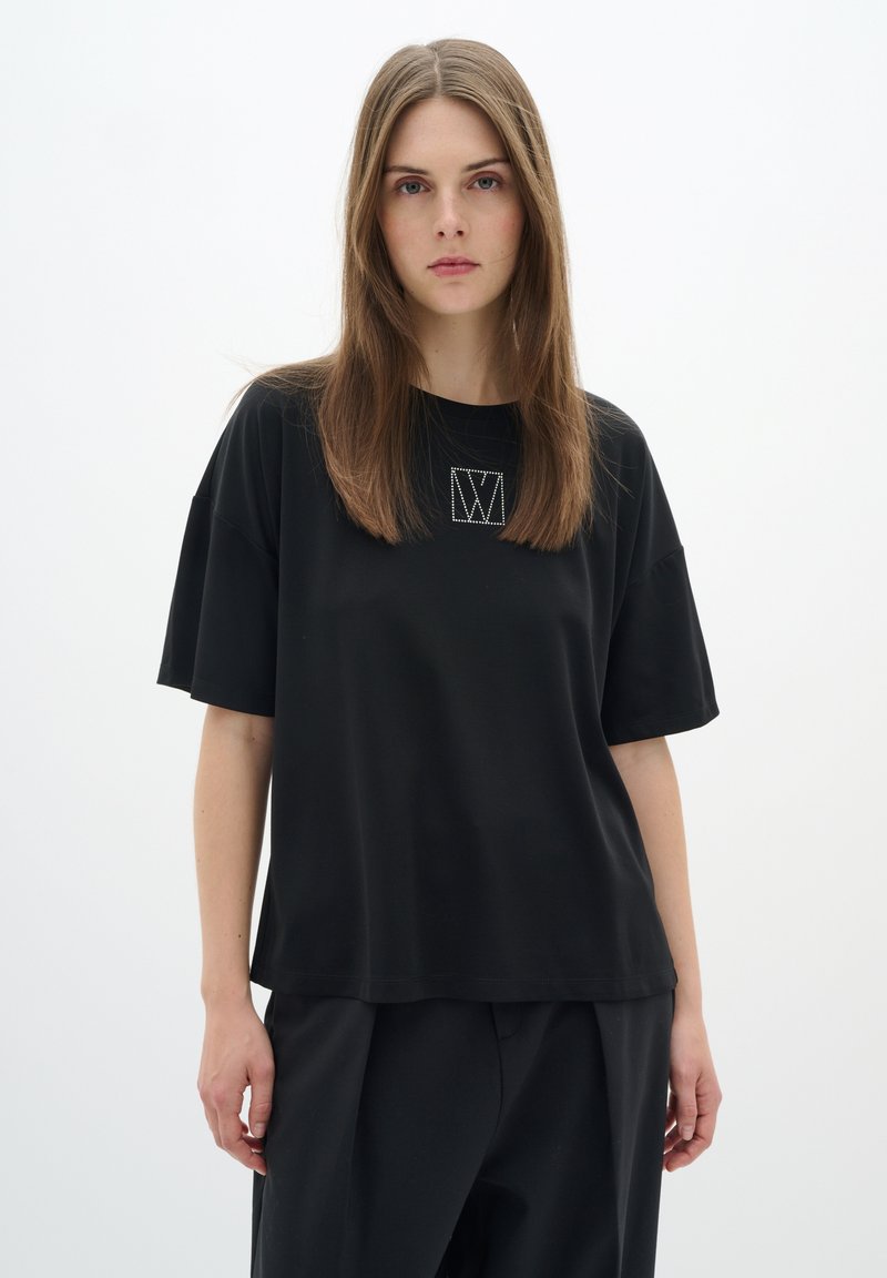 Čierne oversized tričko s krátkymi rukávmi, s vyšitým logom v štvorci na prednej strane. Hladká tkanina a voľný strih.