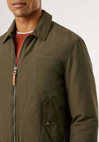 Cazadora bomber verde oliva con cierre frontal, tirador de cremallera de cuero marrón, sutil logo de "Pepe Jeans" y un bolsillo parche con un detalle de botón.