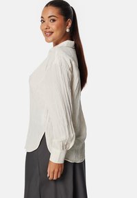 Blouse en tissu froissé de couleur crème, avec des manches longues, un col à boutons, et des fentes sur les côtés, assortie à une jupe sombre.
