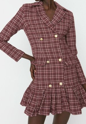 Kobieta w bordowo-białej kratkowanej dwurzędowej sukience marynarkowej z złotymi guzikami i plisowanym dołem, ręka na biodrze.