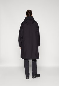 Cappotto lungo nero con cappuccio, realizzato in un tessuto morbido e un design minimalista. Abbinato a pantaloni scuri e calzature robuste.