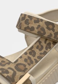 Sandale plateforme à imprimé léopard avec des brides beige et une finition texturée. Présente un accent doré sur la bride pour plus de détails.