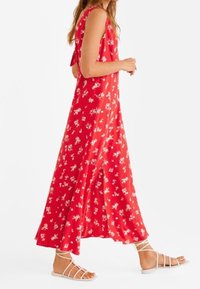 Robe longue en tissu rouge avec motif floral blanc. Présente une coupe sans manches, une coupe ample, et un détail noué à l'arrière. Portée avec des sandales blanches.