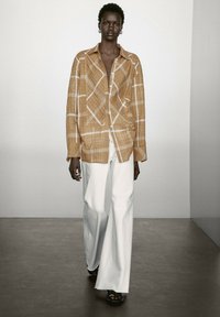 Langarmshirt met een ruitpatroon in warm bruin met witte lijnen, gecombineerd met witte wijde broek en zwarte sandalen op een gladde vloer.