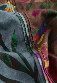 Close-up van stofplooien met kleurrijke bloemmotieven in bruine en grijstinten, met gedetailleerd stikwerk langs de naden.