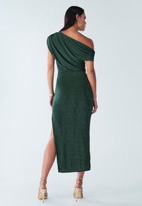 Robe verte asymétrique à une épaule avec un design drapé, tissu texturé et une fente latérale, associée à des talons dorés à lanières.