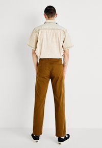 Beige short-sleeve shirt with decorative stitching, paired with brown pants. The outfit features a relaxed fit and minimalistic design.