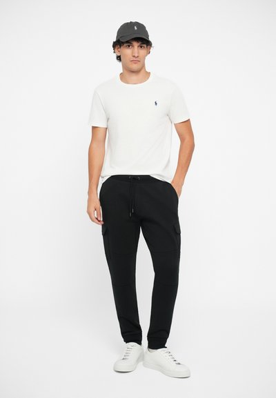 Pantaloni cargo negri cu buzunare laterale, tricou alb cu mânecă scurtă și șapcă gri, asortate cu adidași albi. Ținută simplă, casual.