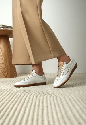 Persoon draagt beige wijde broeken en witte leren sneakers met bruine zolen, staand op een textuur van crème tapijt bij een houten tafel.