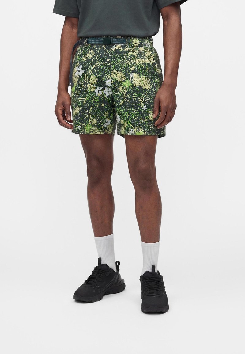 Άνδρας που φορά πράσινα και μαύρα σορτς με λουλουδάτο μοτίβο, μαύρα αθλητικά παπούτσια, λευκές κάλτσες και ένα σκούρο πράσινο μπλουζάκι, στέκεται μπροστά σε λευκό φόντο.