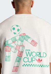 Persona con maglietta bianca con figura pixelata rossa e verde che colpisce di testa un pallone da calcio, testo FIFA World Cup e logo Adidas sul retro.