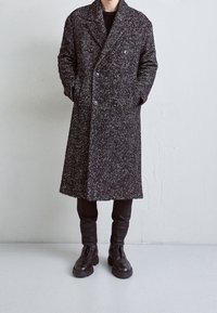 Černobílý vlněný kabát s rybí kostí, dvojřadý a s velkými knoflíky, nošený přes úzké černé kalhoty a boty.