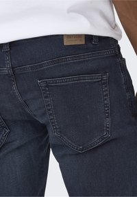 Jeans de mezclilla azul oscuro con bolsillos traseros, que presentan una etiqueta en la cintura y una textura suave. Las costuras son visibles a lo largo de las uniones.