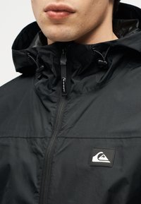 Κοντινό πλάνο ενός ατόμου που φοράει μαύρο μπουφάν με κουκούλα, με φερμουάρ μπροστά και λευκό λογότυπο βουνού στο στήθος.