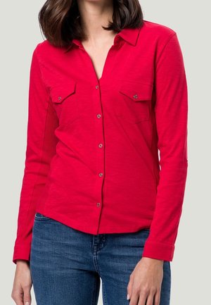 Kobieta ubrana w długą, jasnoczerwoną koszulę na guziki z kieszeniami na piersi oraz niebieskie jeansy, stojąca na jednolitym jasnym tle.